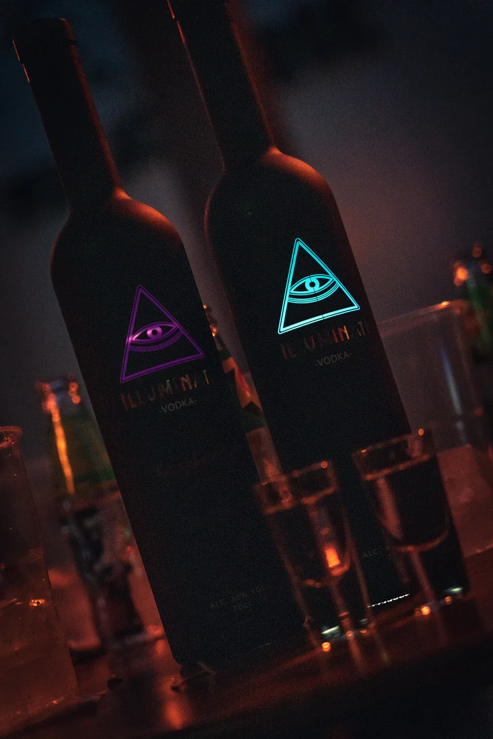 Two Illuminati Vodka bottles in low light, the Raspberry eye glowing pink and the Original eye glowing blue